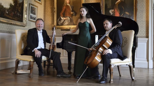 A woman in a green evening dress standing in front of a grand piano. Two men sit either side of her, in evening dress, one holds a cello and the other holds a violin. They are in a country house with oil paintings hanging on the wall behind them.